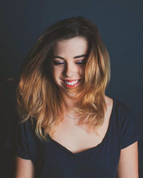 Miranda Sita smiling and looking down, her long ombre hair falling past her shoulders, in a navy T-shirt against a dark blue backdrop.